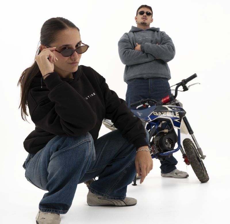 Chica en cuclillas con sudadera negra y chico de pie con sudadera gris junto a una minimoto