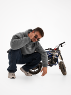 Hombre agachado junto a una minimoto con la sudadera de moto gris washed, bajándose las gafas de sol
