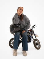 Mujer sentada en una minimoto azul con la sudadera de moto gris washed y un casco de carbono sobre la rodilla